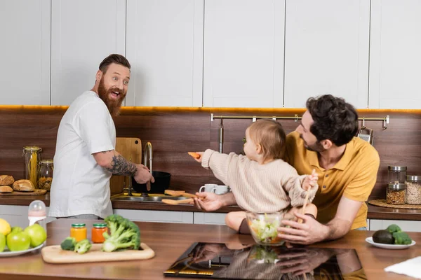 Захоплений гей чоловік дивиться на дочку з овочами поруч з партнером і свіжим салатом на кухні — стокове фото