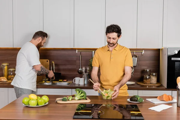 Gay homem cozinhar salada perto de parceiro e comida no cozinha — Fotografia de Stock