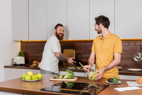 Улыбающаяся бородатая гей-пара готовит дома на кухне — стоковое фото