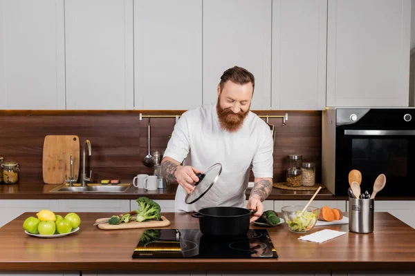Hombre tatuado sosteniendo la tapa cerca de la olla en la estufa y la comida en la cocina - foto de stock