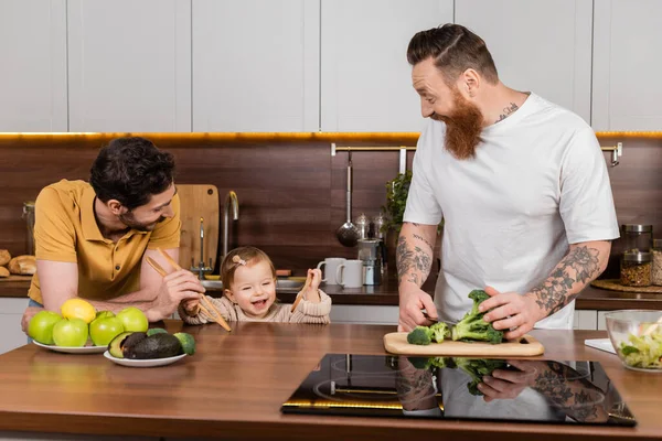 Усміхнена дівчинка-малюк тримає ложки біля батьків, готуючи на кухні — стокове фото