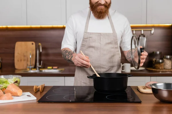 Vista cortada de homem barbudo cozinhar em panela perto de salada na bancada na cozinha — Fotografia de Stock