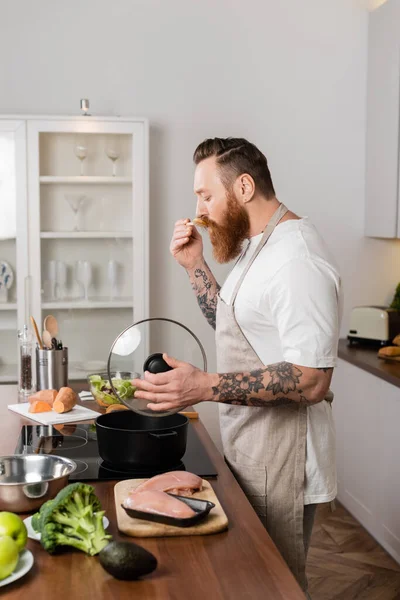 Side view of tattooed man in apron tasting food while cooking in kitchen — Stock Photo