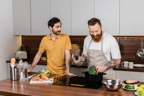 Татуйований гей чоловік кладе броколі в горщик під час приготування їжі біля партнера на кухні — стокове фото