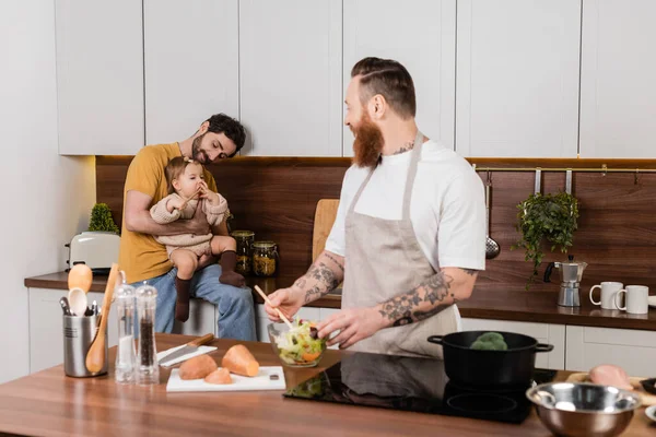 Gay father holding baby daughter while partner cooking in kitchen — Foto stock