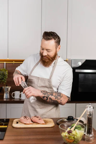 Hombre barbudo y tatuado salando filete de pollo cerca de ensalada fresca en la cocina — Stock Photo
