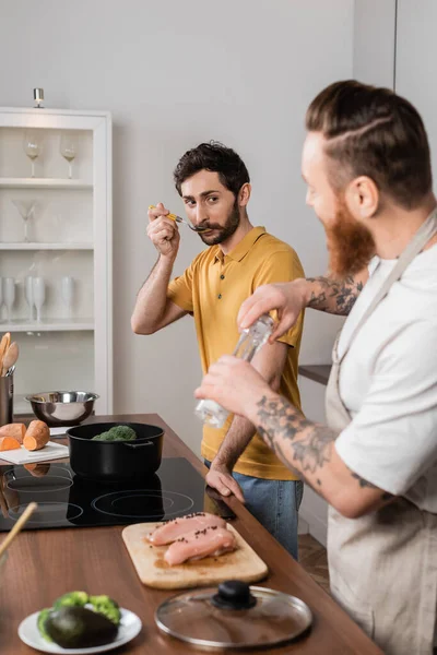 Gay man tasting food while partner salting chicken fillet in kitchen — Stock Photo