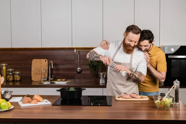 Gay man hugging tattooed husband salting chicken fillet near fresh salad in kitchen - foto de stock