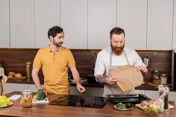 Gay homem segurando assar papel perto cru frango filé e parceiro no cozinha — Fotografia de Stock