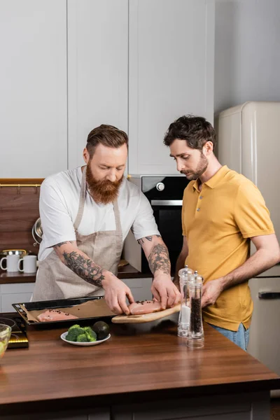 Gay couple cooking chicken fillet near salt and pepper mills in kitchen - foto de stock