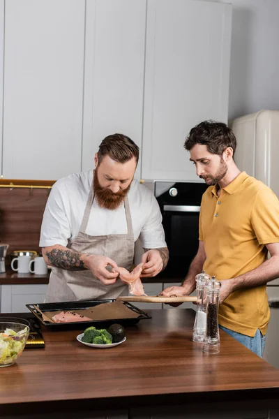 Gay couple putting chicken fillet on baking sheet while cooking in kitchen — Stock Photo