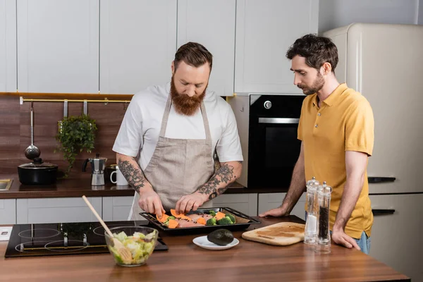Gay man putting vegetables and chicken fillet on baking sheet near partner in kitchen — Stockfoto