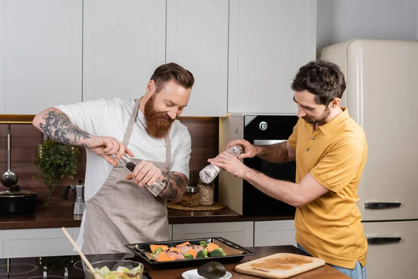 Smiling gay couple seasoning chicken fillet and vegetables on baking sheet in kitchen - foto de stock