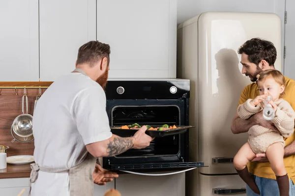 Gay homem colocando cru refeição no forno perto parceiro segurando filha no cozinha — Fotografia de Stock