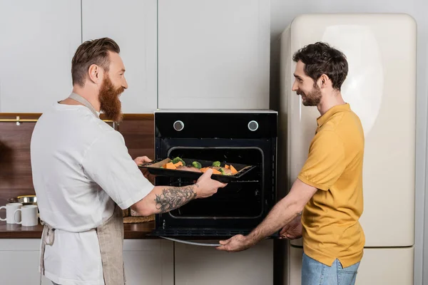 Vista lateral de pareja gay poniendo carne y verduras en bandeja para hornear en el horno en casa - foto de stock