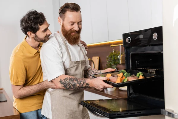 Gay man hugging partner putting chicken fillet and vegetables in oven in kitchen - foto de stock