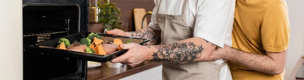 Cropped view of gay man hugging husband putting food in oven while cooking in kitchen, banner — Photo de stock