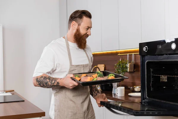 Усміхнений татуйований чоловік тримає лист для випічки з м'ясом та овочами біля печі на кухні — стокове фото