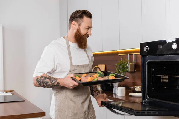 Smiling tattooed man holding baking sheet with meat and vegetables near oven in kitchen — Foto stock