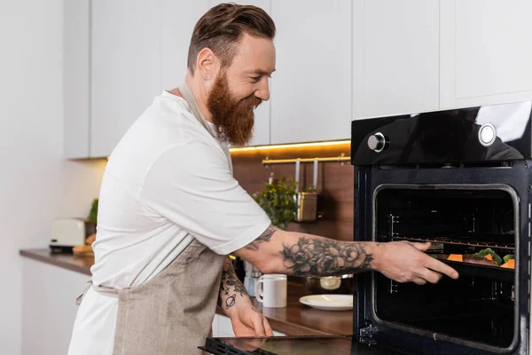 Sorrindo tatuado homem colocando comida no forno enquanto cozinha em casa — Fotografia de Stock