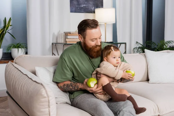 Barbuto padre in possesso di mela e figlia bambino sul divano a casa — Foto stock