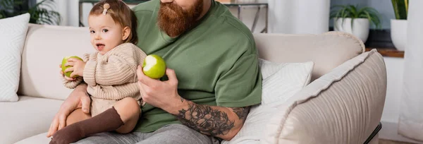 Tattooed father holding fresh apple and toddler daughter in living room, banner — Photo de stock