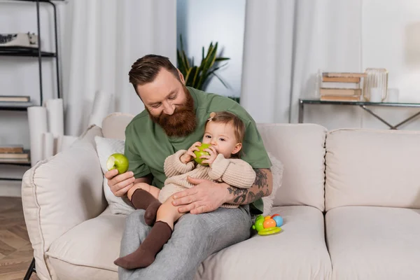 Позитивний бородатий тато тримає яблуко і дивиться на дочку малюка на дивані — стокове фото