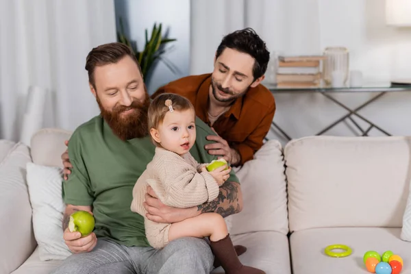 Sonriente pareja gay mirando bebé hija con manzana en sofá en casa - foto de stock
