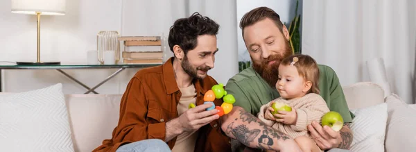 Smiling gay man holding toy near daughter and partner with apples, banner - foto de stock