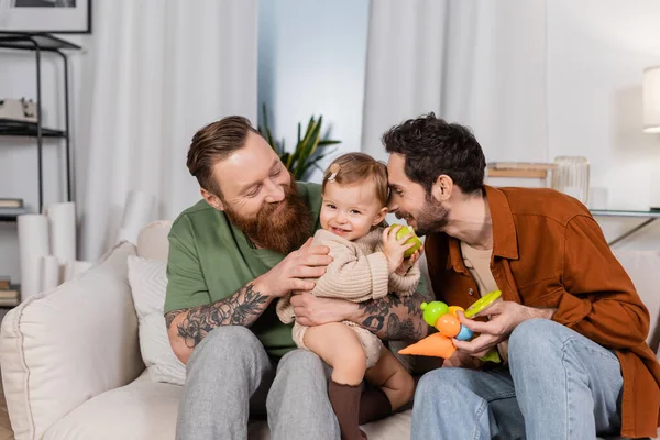 Happy gay couple holding baby girl with apple in living room - foto de stock