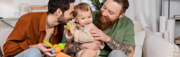 Positive same sex parents looking at baby daughter with apple at home, banner — стоковое фото
