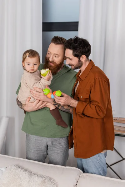 Parents de même sexe tenant jouet et bébé fille avec pomme à la maison — Photo de stock