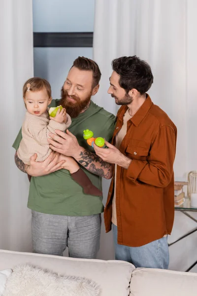 Cheerful gay parents holding baby girl with apple and toy at home — Stock Photo
