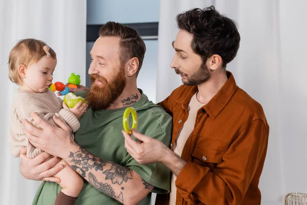 Lächelndes homosexuelles Paar hält Spielzeug und Mädchen mit Apfel zu Hause — Stockfoto