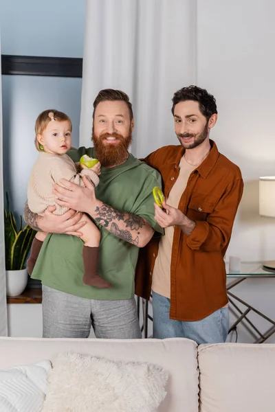 Positive gay man holding baby daughter and looking at camera at home - foto de stock
