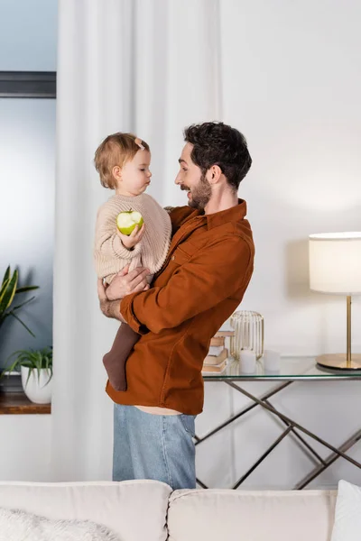 Vue latérale du père joyeux tenant bébé fille avec pomme dans le salon — Photo de stock