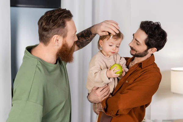 Same sex parents holding baby daughter with apple in living room — стоковое фото