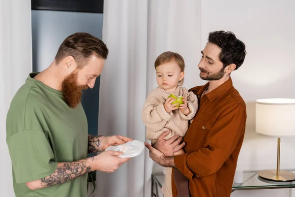 Gay man holding diaper near partner with baby daughter and apple at home - foto de stock