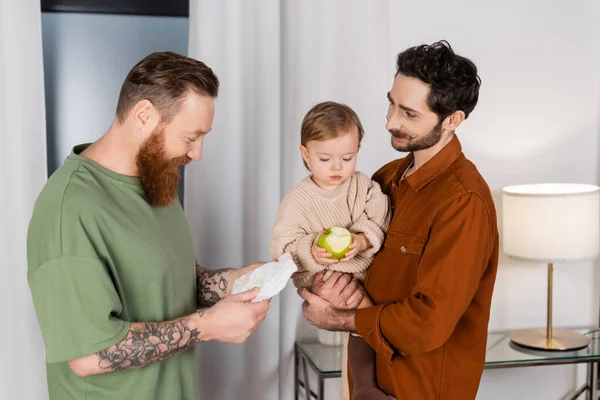 Positive gay couple holding baby daughter and diaper at home - foto de stock