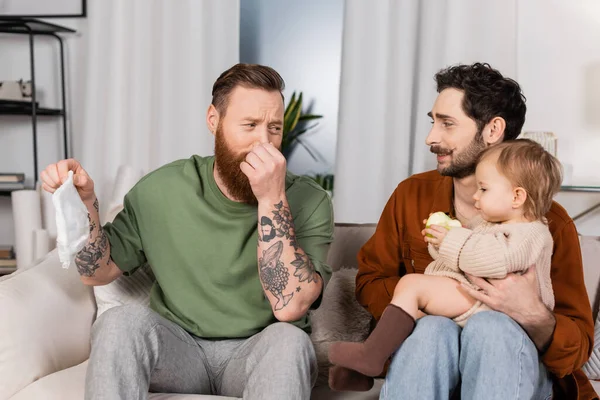 Disgusted gay man plugging nose while holding diaper near partner with baby daughter at home - foto de stock