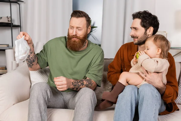 Disgusted gay parents holding diaper and baby girl at home — Stockfoto