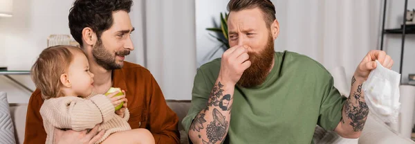 Disgusted gay dad holding diaper near baby daughter with apple and partner at home, banner — Stock Photo