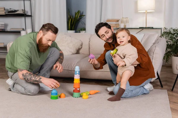 Smiling gay couple playing with baby daughter holding apple at home - foto de stock