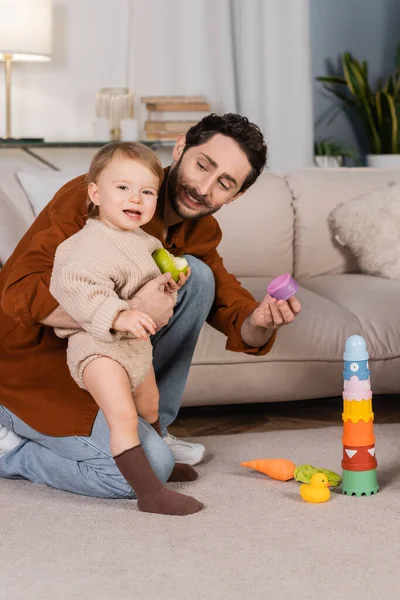 Sorrindo pai segurando brinquedo perto de bebê filha com maçã em casa — Fotografia de Stock