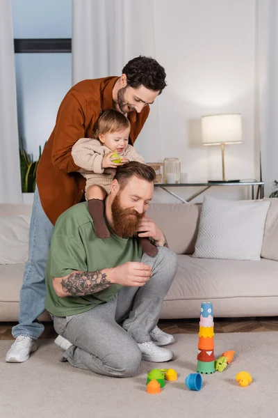 Smiling gay couple playing with baby girl holding apple at home — Stock Photo