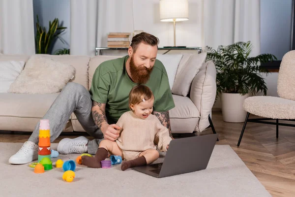 Tattooed father looking at laptop near daughter and toys at home — Photo de stock