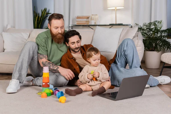 Gay pareja buscando en portátil cerca bebé hija con juguetes en casa - foto de stock