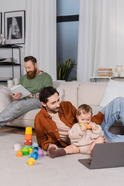 Gay genitore seduta vicino bambino figlia con giocattolo e laptop in soggiorno — Foto stock