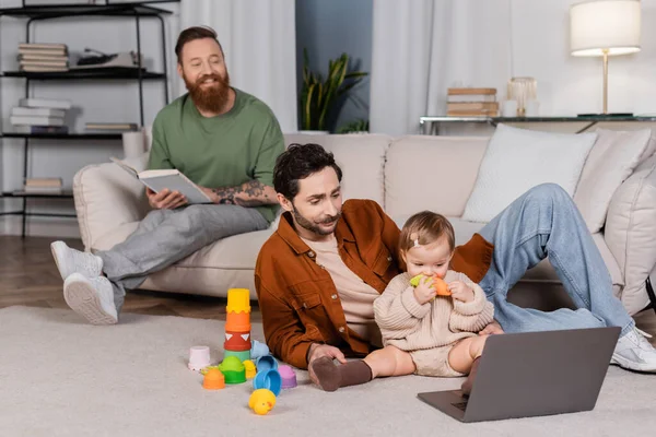 Bebé hija sosteniendo juguete cerca de la computadora portátil y los padres en casa - foto de stock