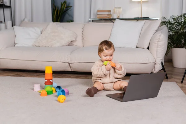 Bebé niña sosteniendo juguete cerca de la computadora portátil en el suelo en casa - foto de stock